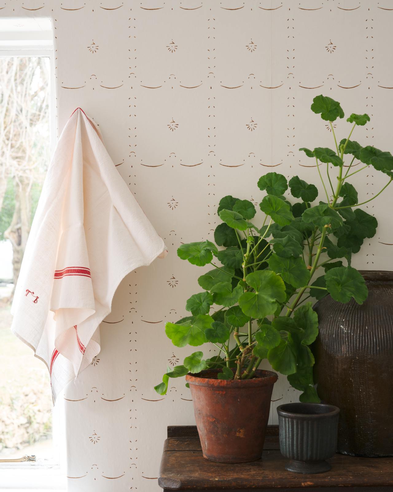 A delicate hand-painted wallpaper around a window with lush geraniums and a vintage tea towel hanging nearby.