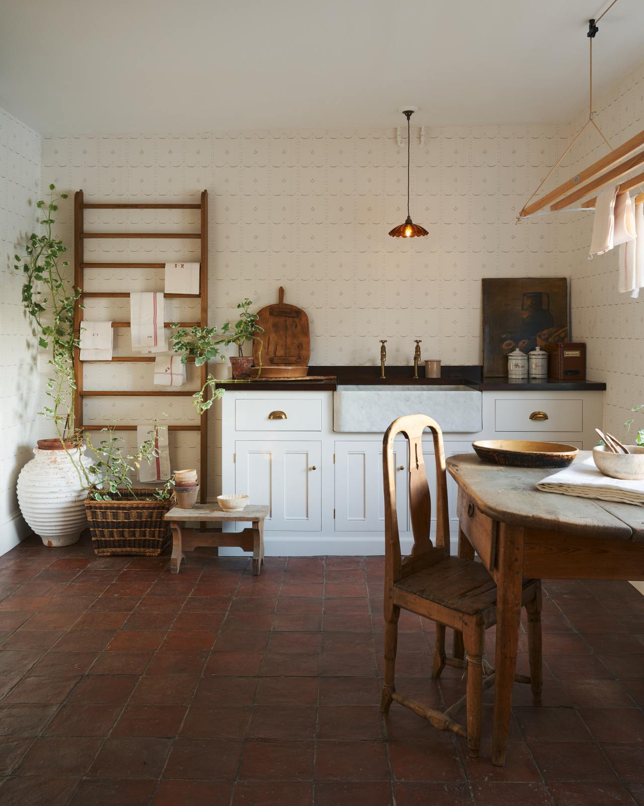 A Mediterranean-inspired scullery with a terracotta floor, white cabinets and a delicate off-white patterned wallpaper.