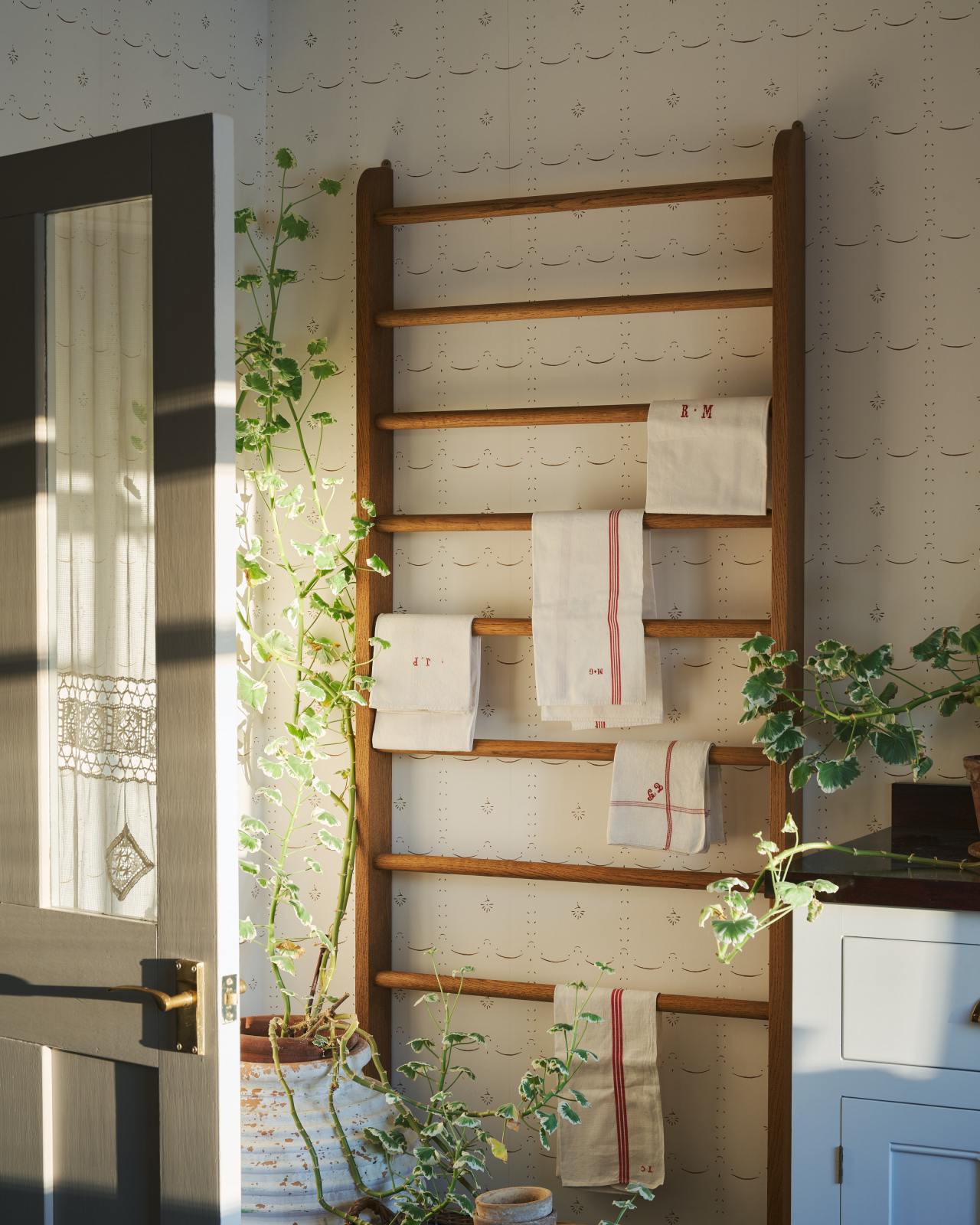 Strong light pours across a delicate patterned wallpaper behind a mid-century-style oak hanging rack which is covered in vintage tea towels.