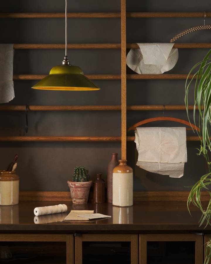 A single chartreuse-coloured pendant light hangs in front of mid-century-style oak racking, the light is on and illuminating the copper worktop below. The light shade is glossy ceramic, shaped like a bonnet hat, with a dark oxidised brass fitting and an oatmeal-coloured fabric cable.