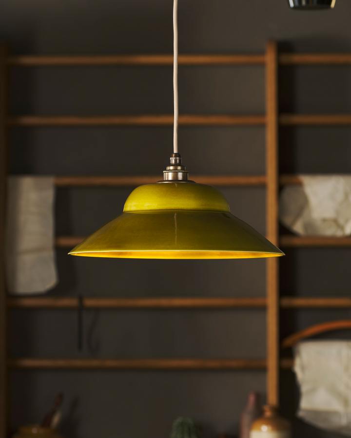 A single chartreuse-coloured pendant light hangs in front of mid-century-style oak racking, the light is on and illuminating the copper worktop below. The light shade is glossy ceramic, shaped like a bonnet hat, with a dark oxidised brass fitting and an oatmeal-coloured fabric cable.