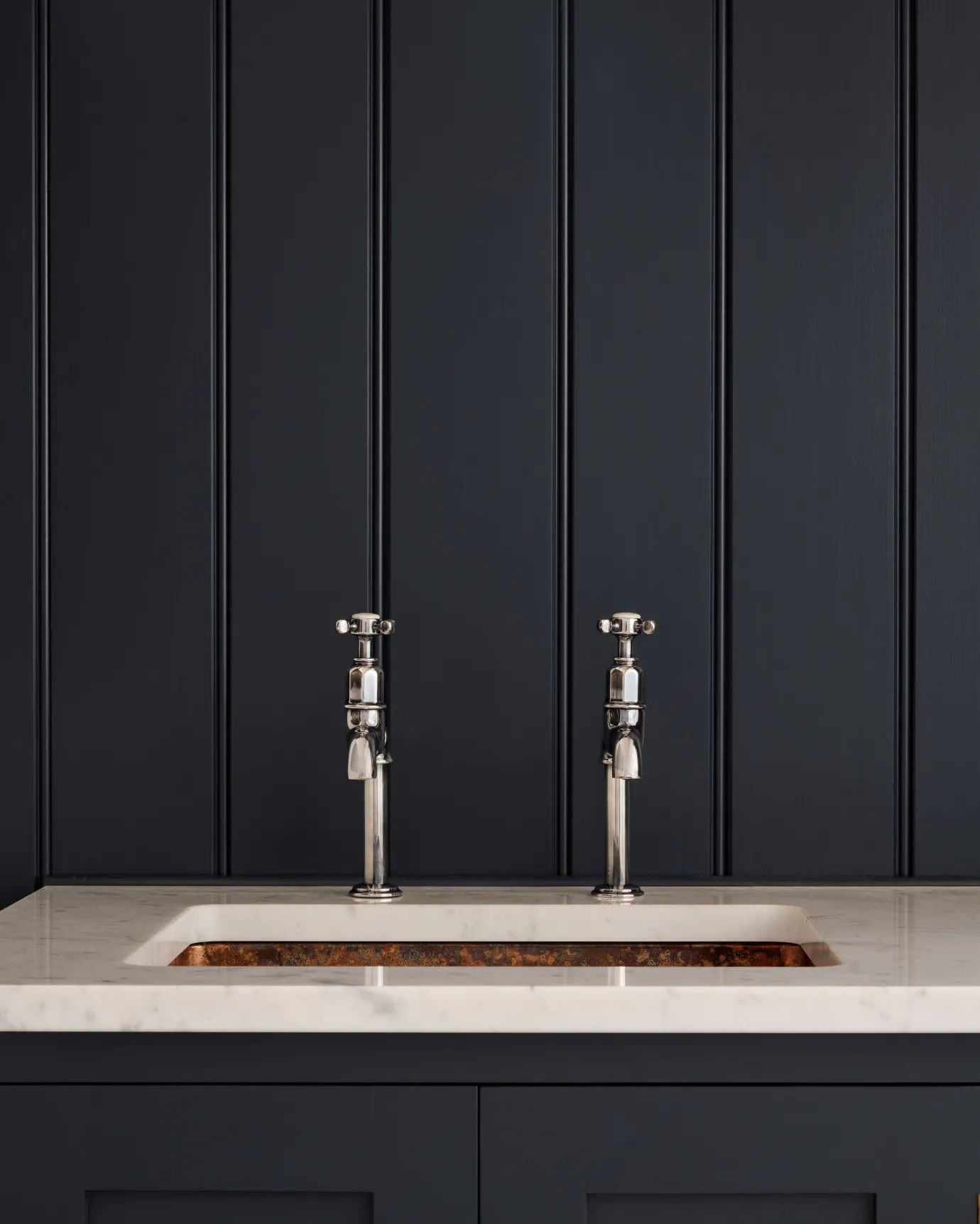 A straight-on view of a pair of traditional-style tarnished silver bibcock taps with crosshead handles in a classic utility kitchen with a Carrara marble worktop and deep blue painted cupboards and tongue and groove panelling.
