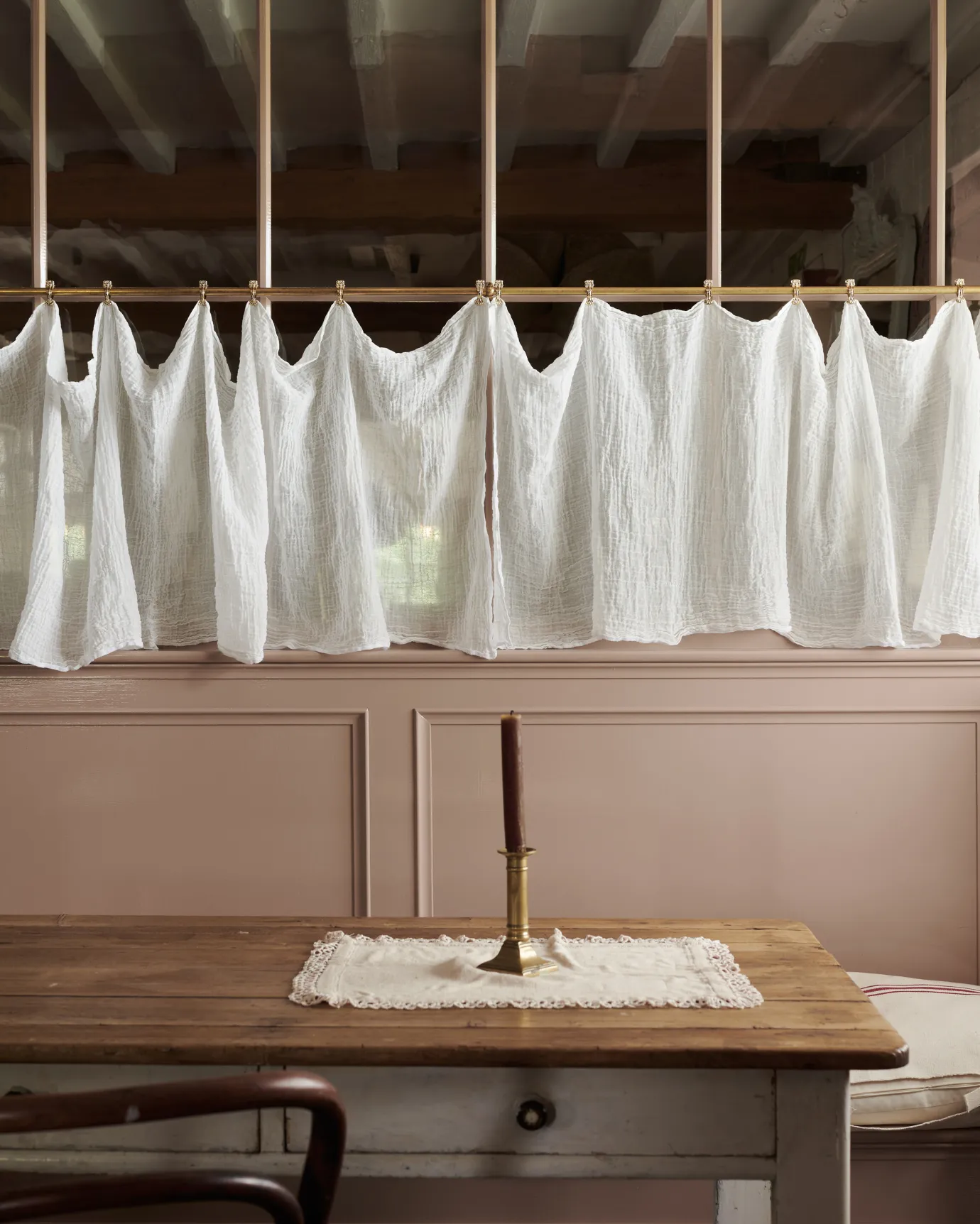 A straight-on view of a traditional pink painted glazed partition wall with an aged brass curtain rail attached to the central horizontal frame. A delicate linen curtain is clipped in several places along the length of the rail.