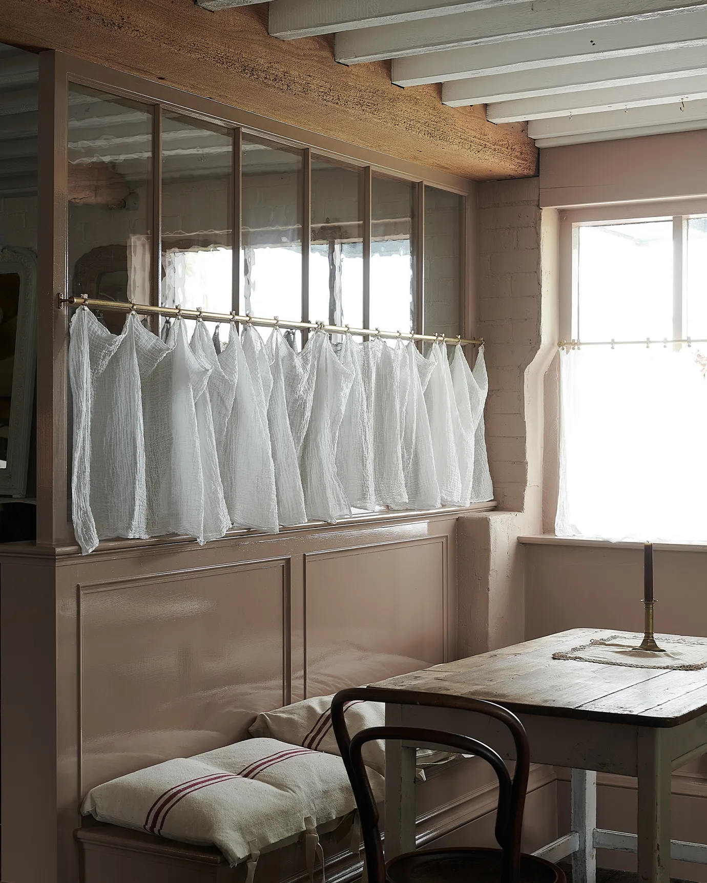 An oblique view of a traditional pink painted glazed partition wall with an aged brass curtain rail attached to the central horizontal frame. A delicate lace curtain is clipped in several places along the length of the rail.